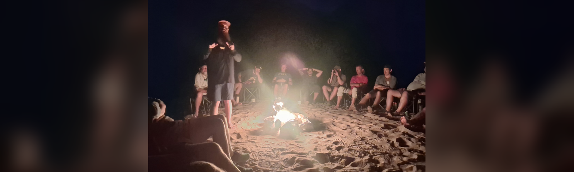 A group of hikers gathering around a campfire as a standing speaker shares with them